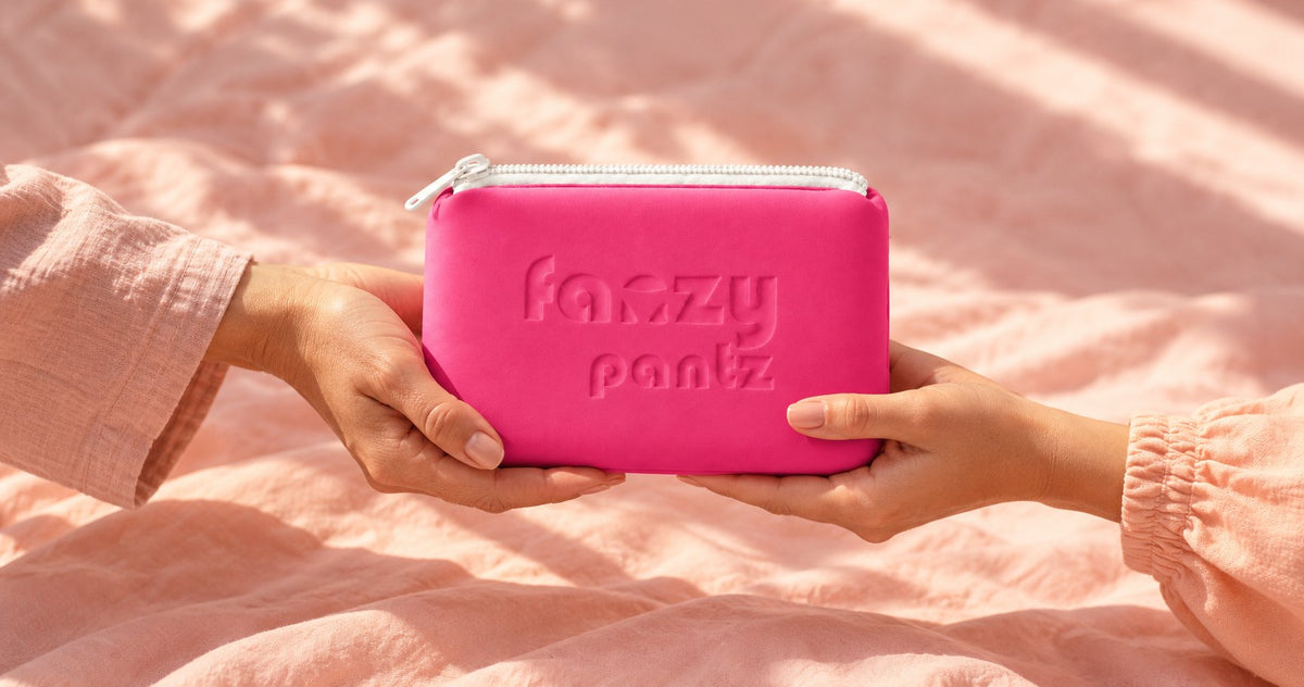 Two hands passing over a pink Fanzy Pantz pouch on pastel bedding